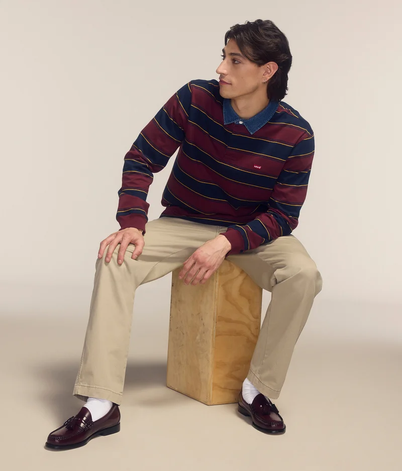 Man sitting on a wooden stool, wearing a red, blue, and yellow striped shirt, and khaki pants.