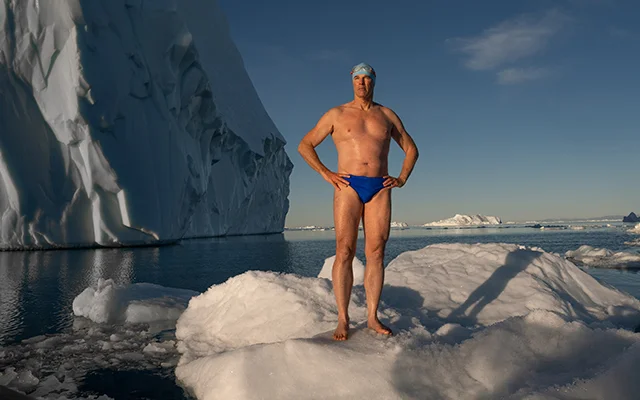 A man stands on a large piece of ice in a swimsuit. There is an ice berg seen behind him.