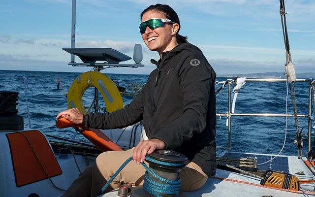 A woman smiles as she steers on a sailboat off the coast. She is wearing sunglasses and the sun is reflected in them. It is a clear and bright day.