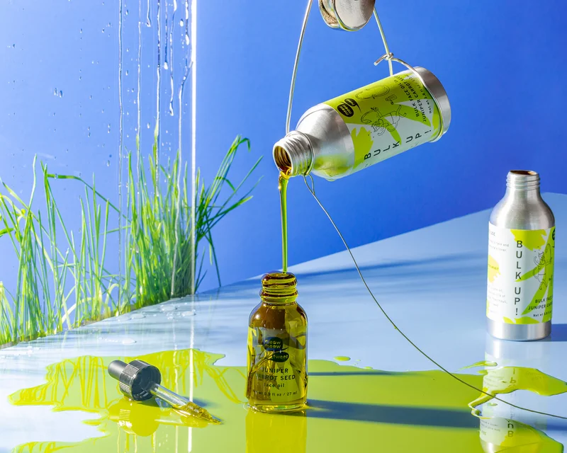A metal bottle pours deep green oil into a smaller glass bottle that overflows. There is an oil puddle on the blue surface.