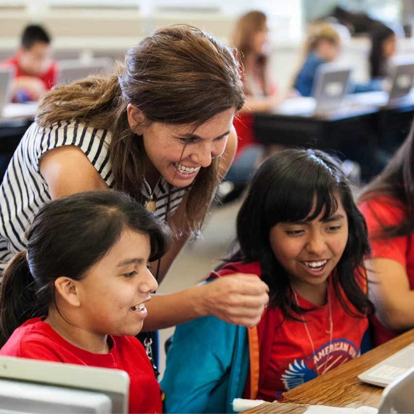 Smiling teacher leaning over to help two students working on a laptop in a classroom.
