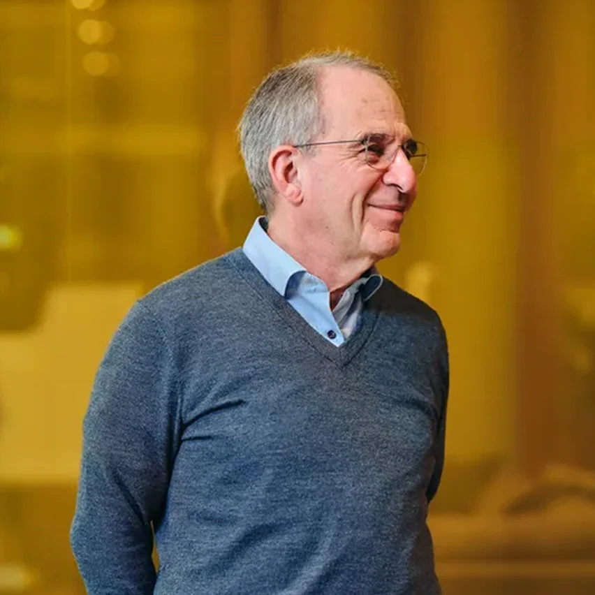 A man with glasses and graying hair, wearing a blue sweater, smiles as he looks to the side.