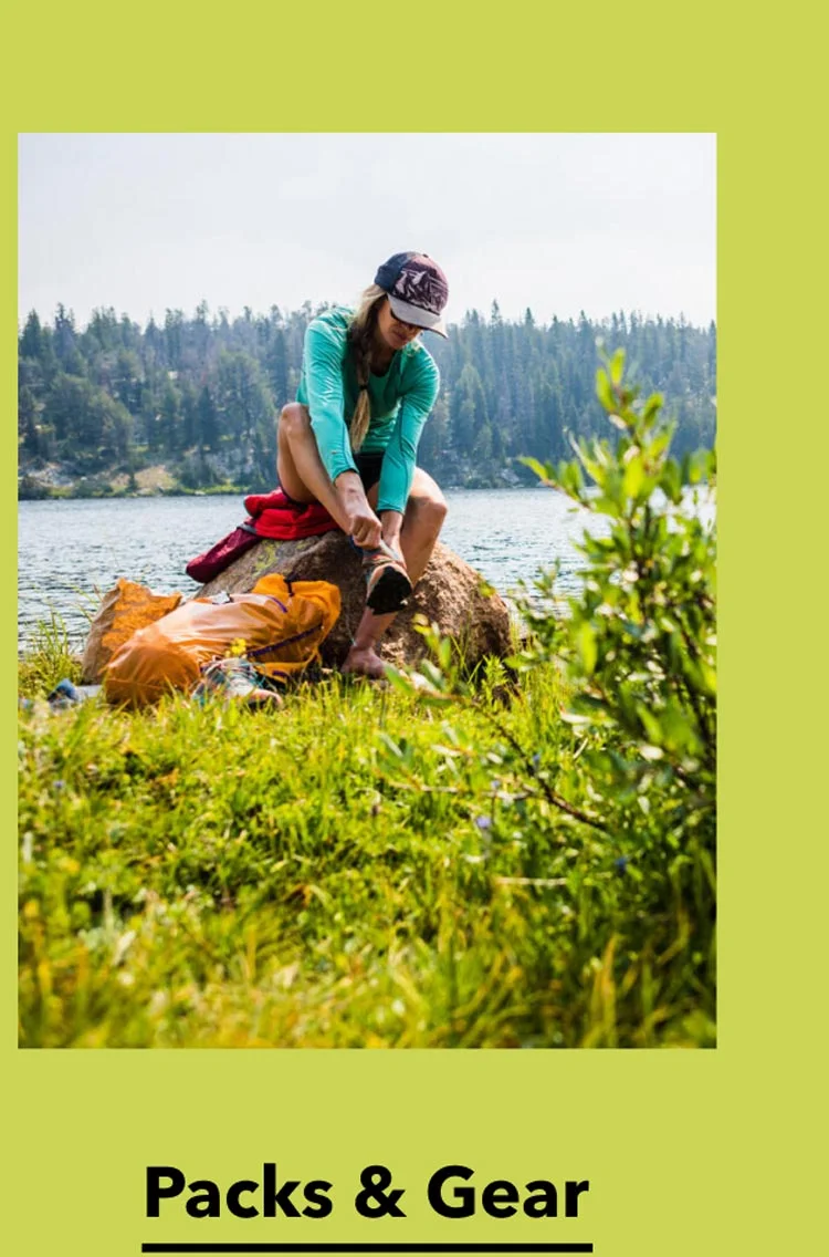 A hiker puts their shoe back on while sitting on a rock beside an alpine lake.