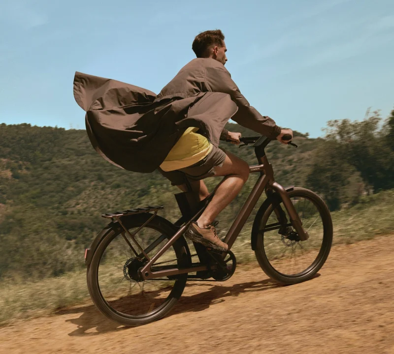 A photograph. A woman riding a Cowboy e-bike through an urban area, with her coat flowing dramatically behind her.