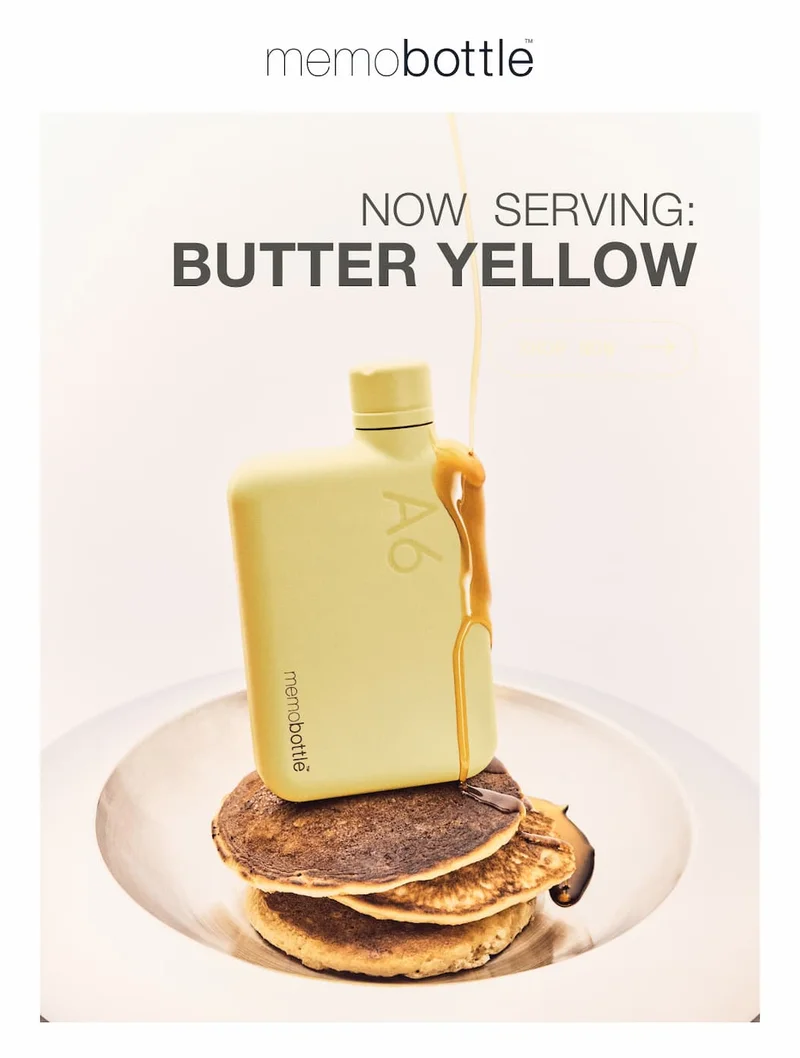 A pale yellow memobottle with the text 'A6' and 'memobottle' printed on it, is placed on top of a stack of pancakes on a silver plate, with honey bein...