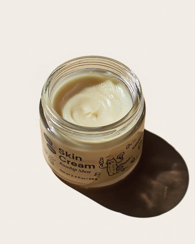 A tan cream in a glass jar sits in a bright light against a cream backdrop