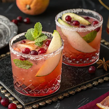 Two cocktail glasses with pink drinks, garnished with mint, cranberries, and apple slices.
