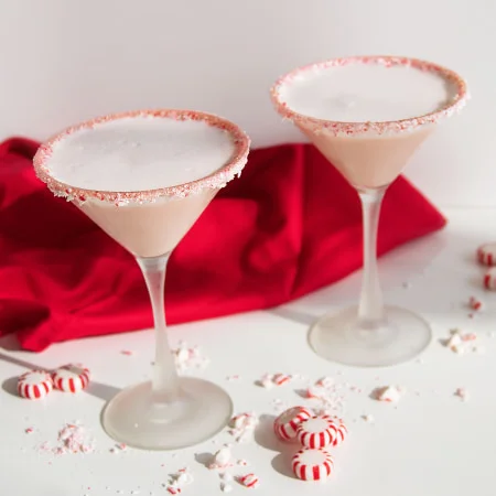 Two cocktail glasses with frosted rims covered in crushed candy, filled with a creamy white drink, surrounded by peppermint candies on a white surface.