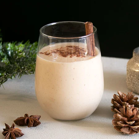 Glass of creamy eggnog with cinnamon stick and sprinkled cinnamon on top, surrounded by fir branches, star anise, and a pinecone.