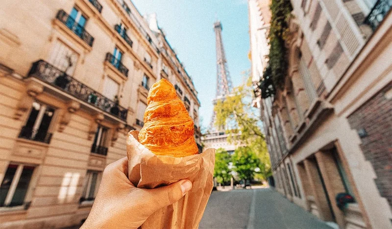 Image description: Woman holding up croissant with view of Eiffel Tower, in Paris