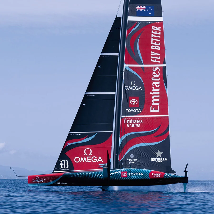A sailboat with Emirates Team New Zealand branding, including OMEGA and other sponsors' logos, sailing on open water.