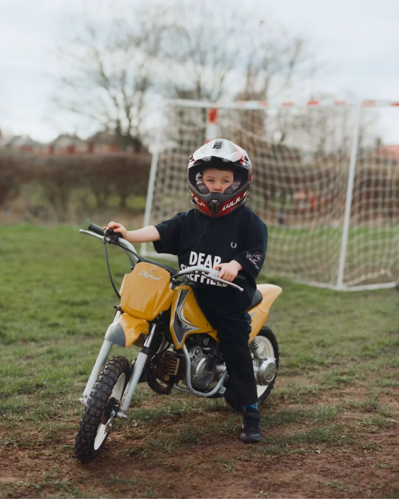 Our Collaborations: small young boy with a helmet on sitting on a child's moped with the Fred Perry x Corbin Shaw 'Dear Sheffield' collaboration polo shirt on.