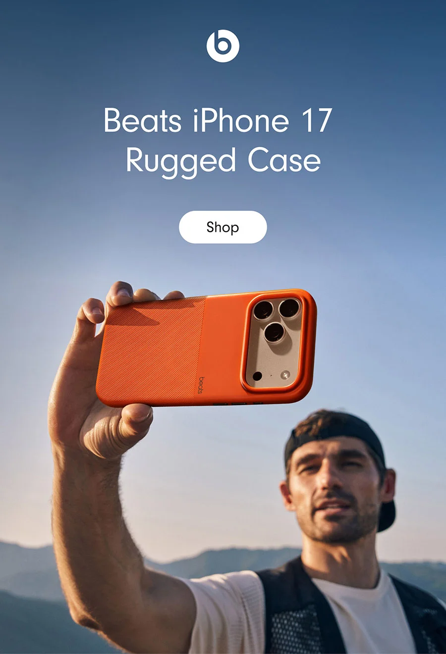 Beats iPhone 17 Rugged Case. A man holding the Beats iPhone 17 Rugged Case with MagSafe and Camera Control in Sierra Orange, against a backdrop of mountains and a clear sky.