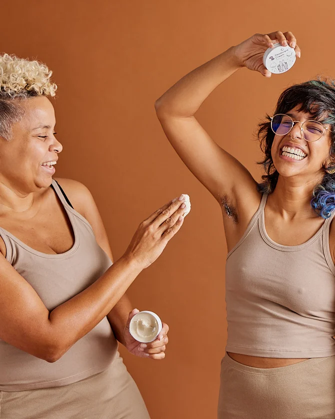 One brown-skinned person applies a huge scoop of cream deodorant to another brown-skinned person's pit. They are laughing.