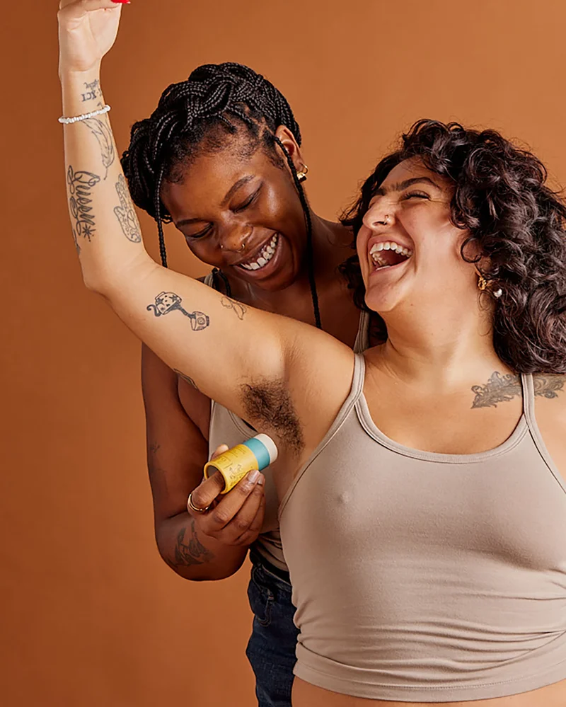 One dark brown skinned person applies a stick deodorant to another brown-skinned person's pit. They are laughing.
