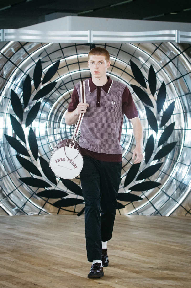 White ginger male model with a buzzcut walks the Fred Perry Runway wearing a tonic waffle knitted shirt in Oxblood and White while carrying a matching barrel bag over his shoulder.