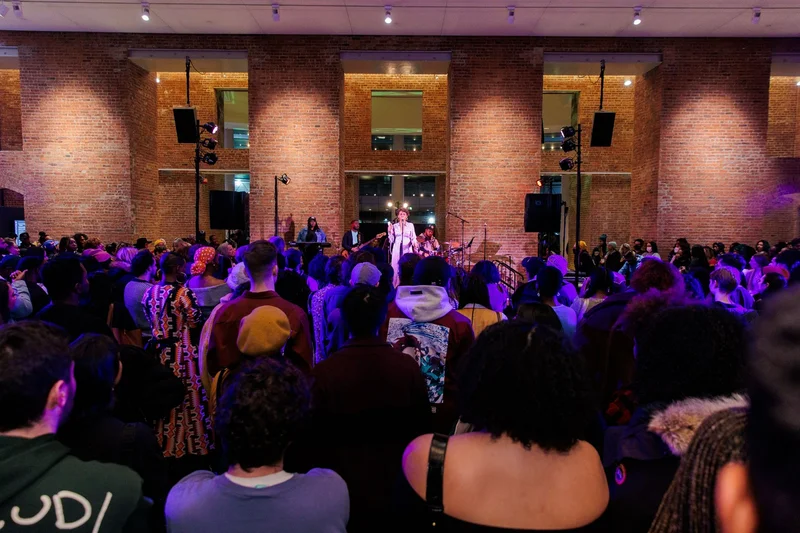 A photograph of people gathered around a stage, where a performer stands at the mic