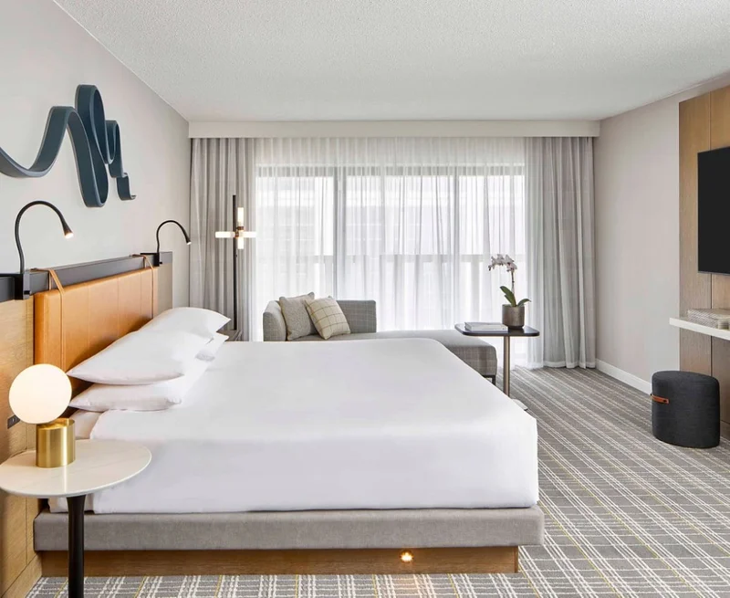 Image description: Hotel room with striped gray and white carpet. A large white bed is in the center of the room and a window with sheer curtains is at the far end.