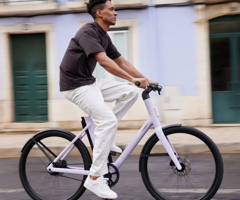A photograph. A man riding a light purple Cowboy e-bike down a city street, with colorful, classic European-style buildings in the background..