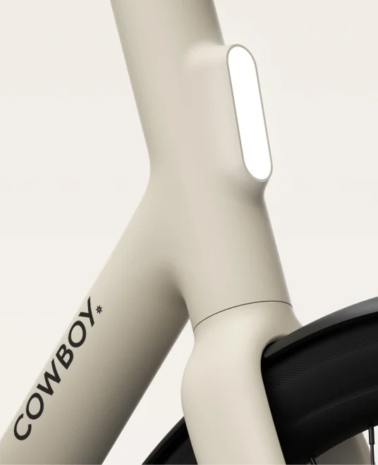 A render. Close-up of a Cowboy e-bike frame in a cream color, featuring a front light integrated into the bike’s sleek design. The Cowboy logo is visible on the frame.