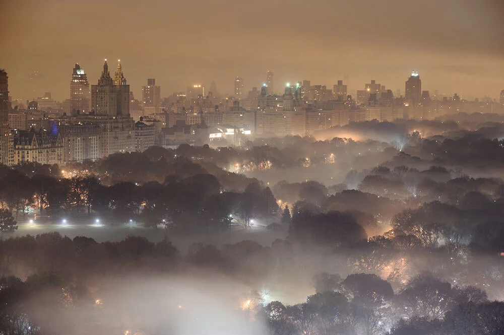 Light pollution and fog combining to blur a New York City skyline at night. Central park sits eerily blanketed by fog in the foreground.