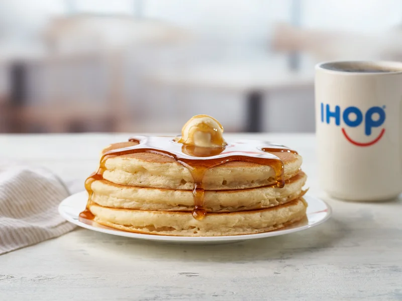 A short stack of IHOP pancakes on a plate, topped with butter and syrup. A mug of coffee is beside it.