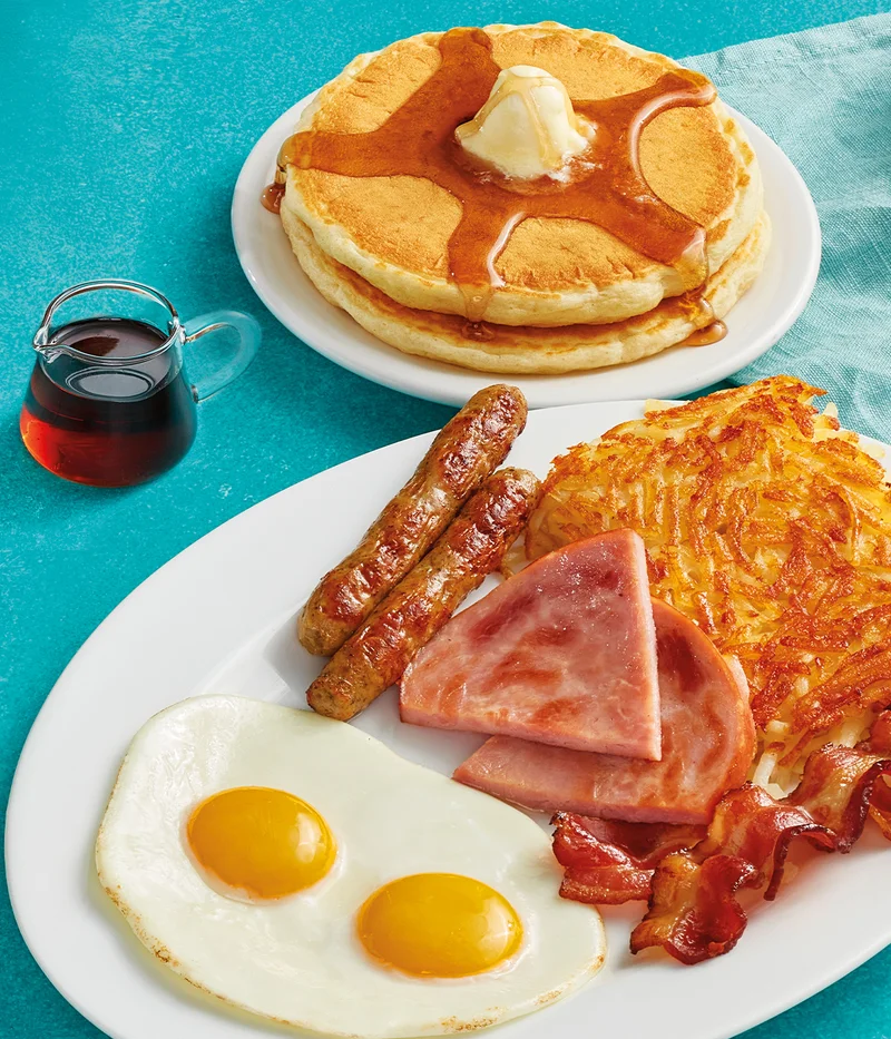Two eggs served your way, two pork sausage links, two slices of thick-cut ham, two strips of hickory-smoked bacon, and hash browns on a plate. Served with 2 buttermilk pancakes topped with syrup and butter.