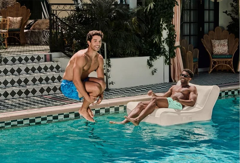 Two men relaxing by a pool in bright swimwear, one jumping in, under headline “The party starter.”