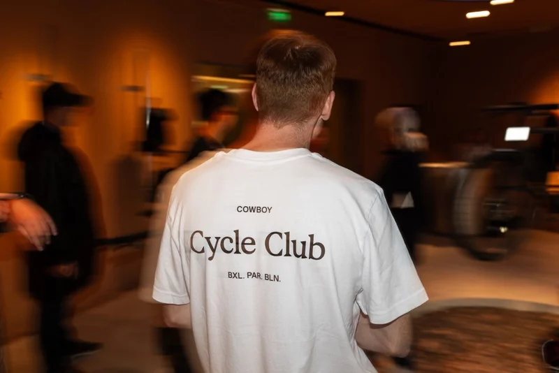 A photograph. A rear-shot of a man wearing a white Cowboy Cycle Club t-shirt