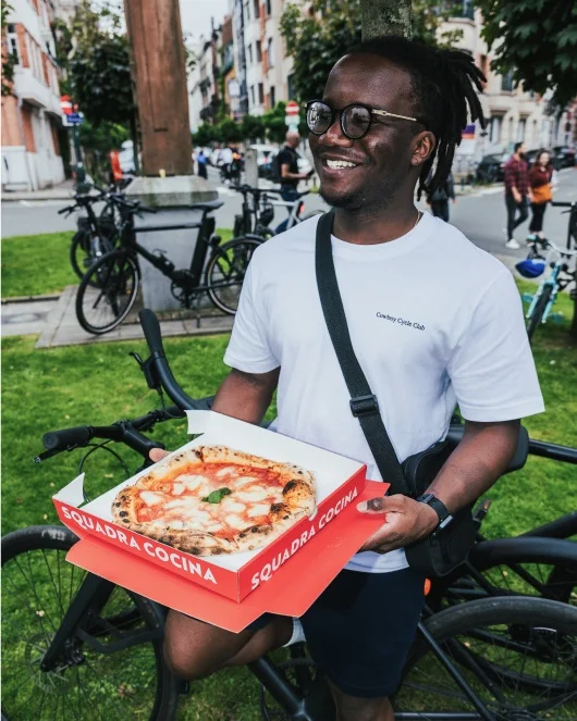 A photograph. A man stands in a green city square holding an open pizza box, several parked Cowboy bikes are visible behind him