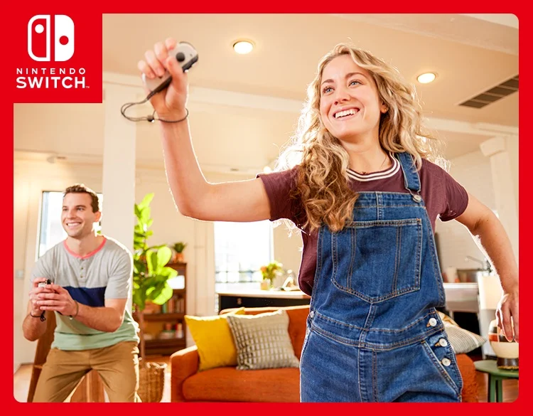 Two people, with big smiles on their faces, are in active poses while playing a Nintendo Switch game using motion controls. Each of them is holding a single Joy-Con Controller.