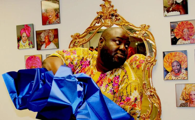 A man in a yellow garment holding blue fabric, in front of a wall featuring pictures of women in headwraps