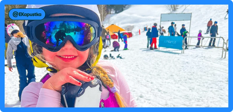 Image Description: Little girl in ski goggles and helmet smiling at the camera with slopes in the background.