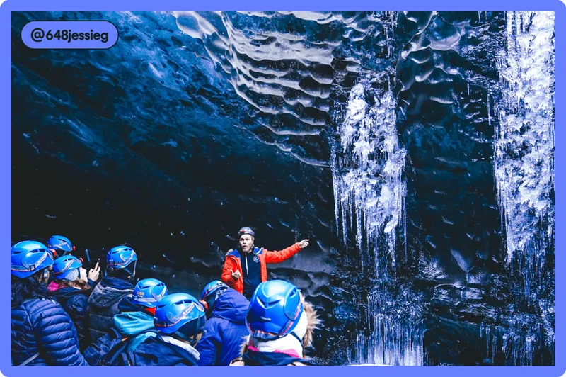 Image Description: Tour group in ice cave with tour guide in a red jacket pointing toward icicles and a large group all in helmets and dark-colored jackets listening to him.