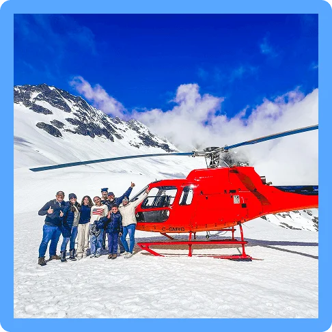 Image Description: Group of people posing next to a red helicopter with snow and mountains behind them.