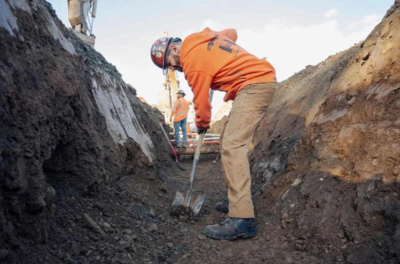A construction worker in an orange hoodie and tan work pants digs with a shovel in a trench, wearing the new Vicious BOA® work boots, designed for durability and comfort in tough work environments.