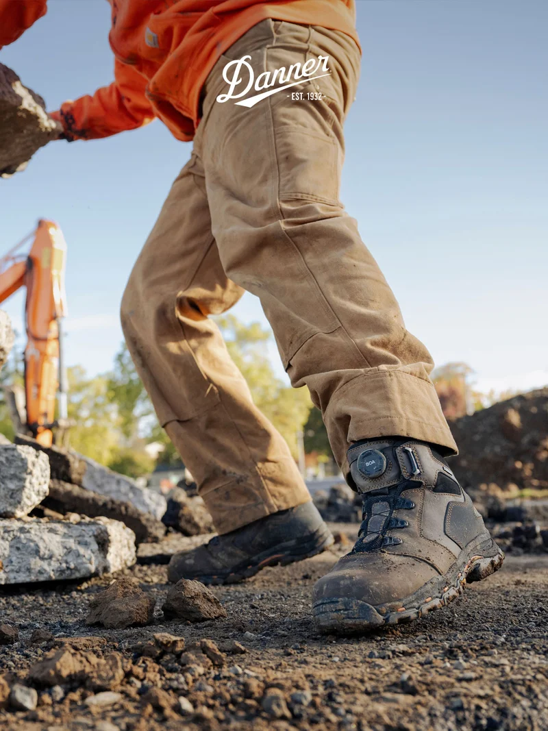 A worker in an orange hoodie and durable work pants navigates a rocky construction site, wearing the new Vicious BOA® work boots equipped with an adjustable dial for a secure fit.