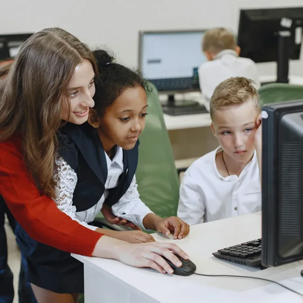 A teacher assists two young students, one girl and one boy, as they look at a computer monitor and use a mouse in a classroom setting.