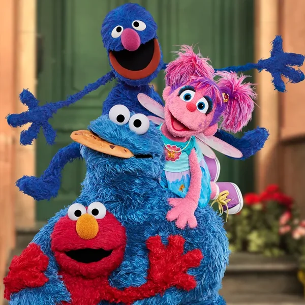 A close-up image showing four Sesame Street Muppets. Cookie Monster, a blue Muppet, is in the center, holding a cookie in his mouth. Seated on his shoulders is Grover, another blue Muppet. Sitting to the right is Abby Cadabby, a pink Muppet fairy. Elmo, a red Muppet, is visible at the bottom of the image.