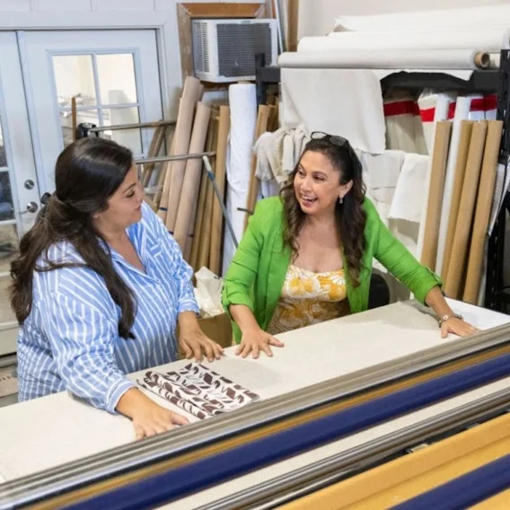 Photo of two women sitting at a desk looking at a piece of fabric and smiling at each other.
