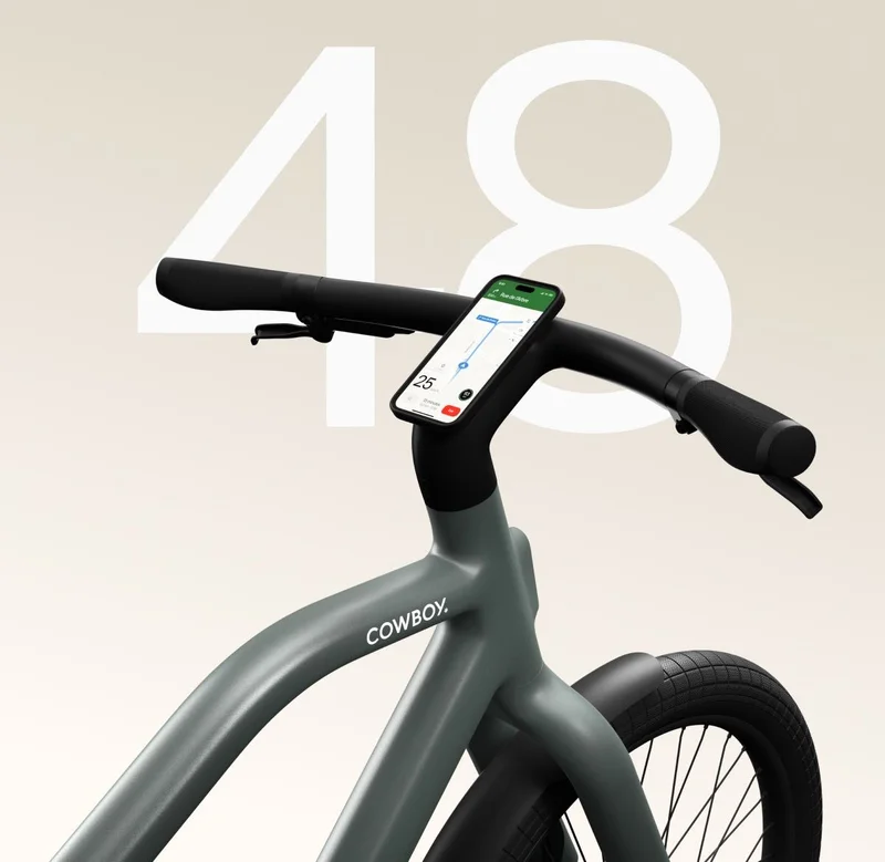 Close-up of the handlebars of a green Cowboy e-bike, with a smartphone mounted on the stem displaying navigation on the screen, and a large number “48” in the background.