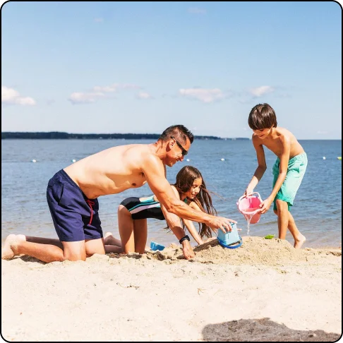 Image Description: A man makes sandcastles with two children on a golden sand beach with water in the background.