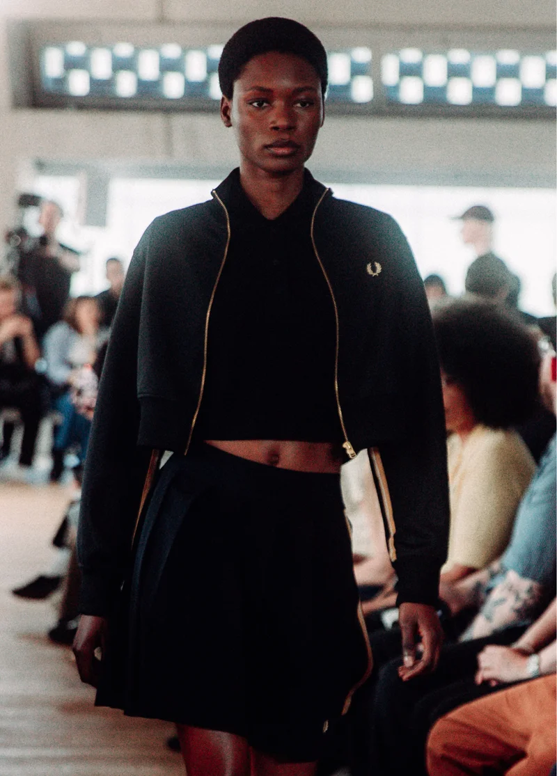 Female black model with short natural textured hair walks our runway wearing a cropped black track jacket with a full gold zip and a black mini skirt with Champagne taping