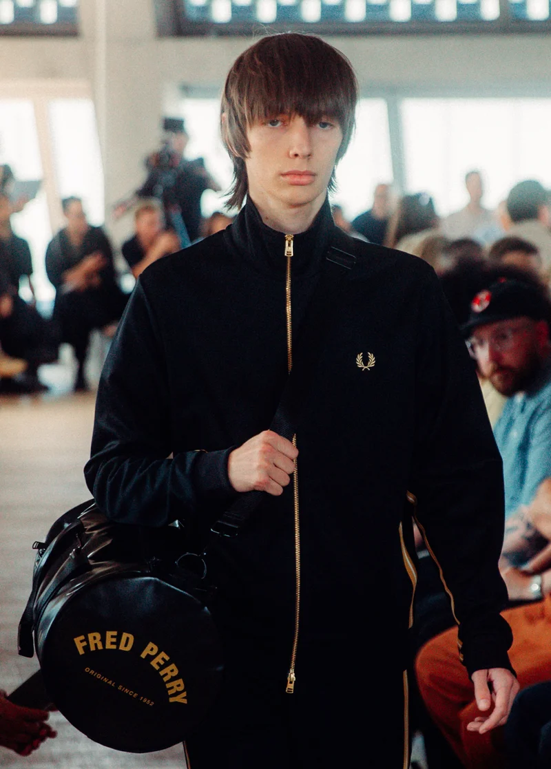 White male model with light brown straight hair walks our runway wearing Black track jacket with Champagne taping and a gold full zip and holds the Black Champagne barrel bag