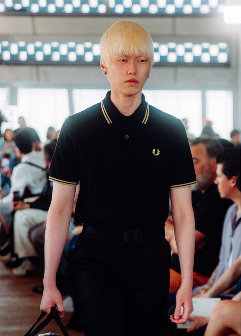 Asian male modern with straight bleached blonde hair walks our runway wearing the twin-tipped Fred Perry shirt in Black / Champagne
