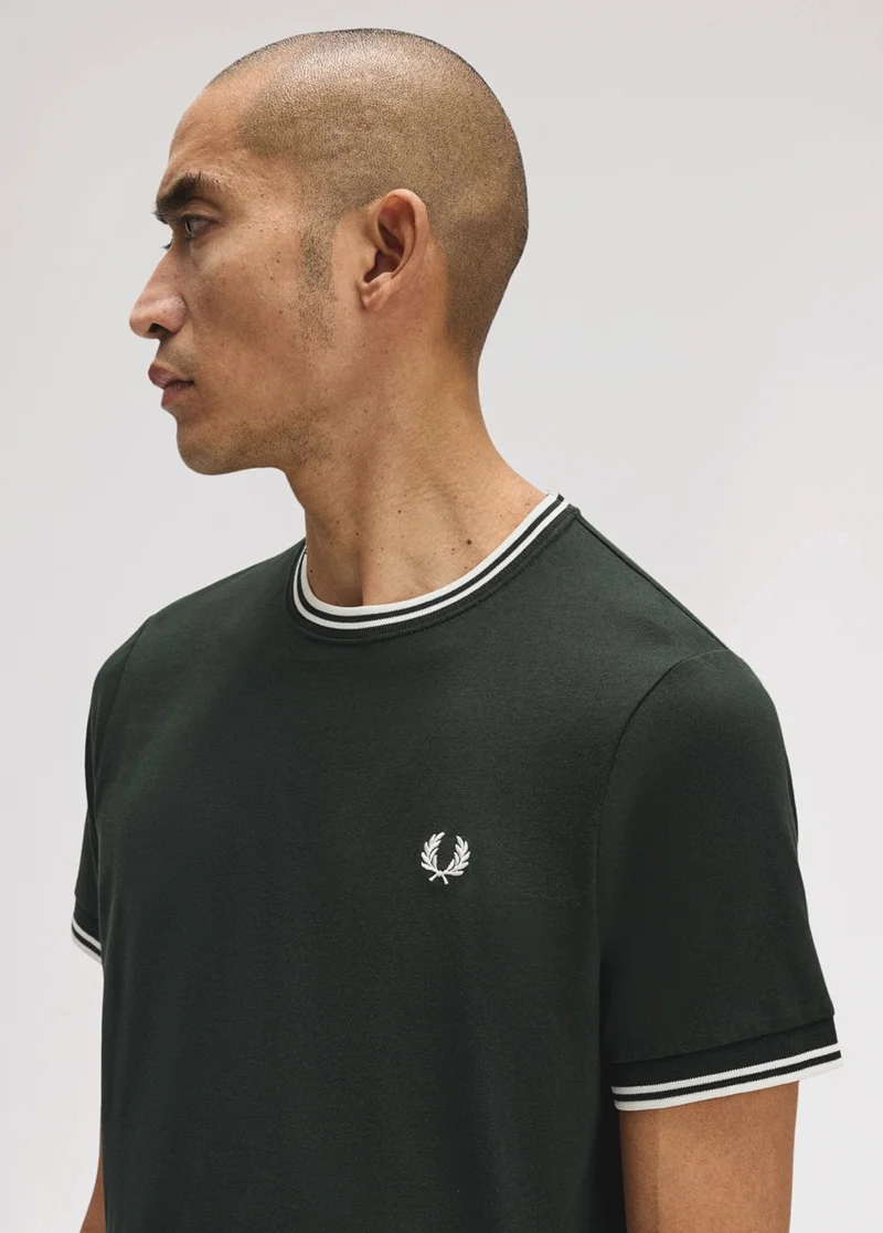 Mixed race male model with shaved head looks to the side nonchalantly while wearing a dark green T-shirt with white twin-tipping on the collar and sleeves