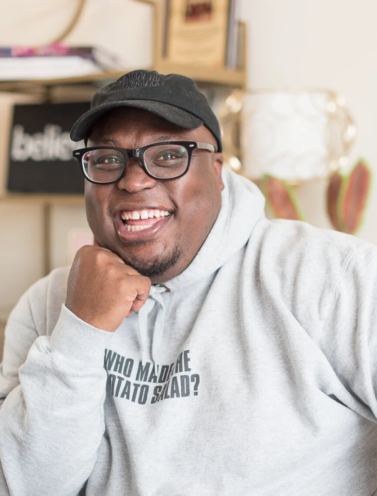 Food historian and community organizer KJ Kearney, wearing glasses and a cap, smiling warmly with his hand under his chin. He’s in a gray hoodie with "Who Made the Potato Salad?" printed on it.