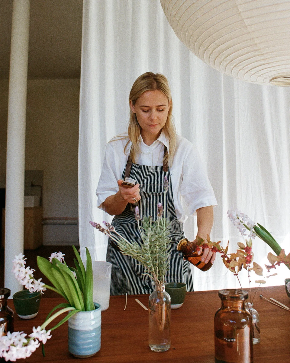 Blonde woman in an apron setting up for a candle-making workshop. Flowers in vases adorn the wooden table in the foreground.