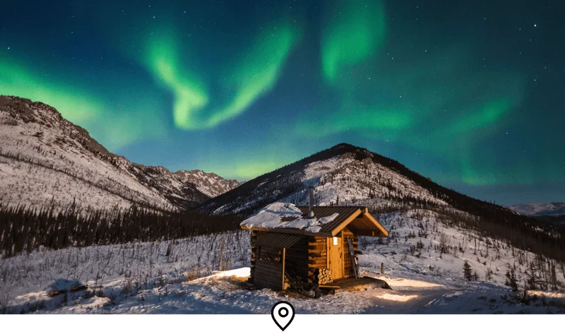 Image Description: Landscape shot of a log cabin in Alaska’s White Mountains National Recreation Area, blanketed by a night sky of aurora lights