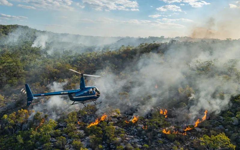 A helicopter flies over a section of a large forest on fire.